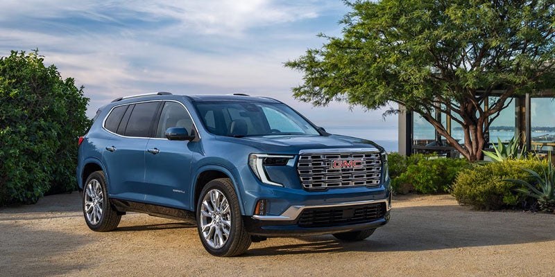 A blue GMC Acadia Denali SUV parked on a gravel driveway in front of a modern building with a large tree.