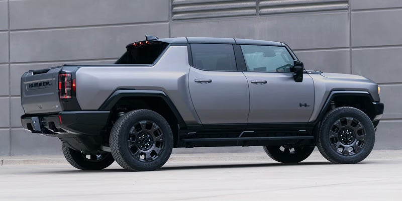 The rear three-quarter view of a gray GMC Hummer EV parked on a paved surface with a gray building behind it.