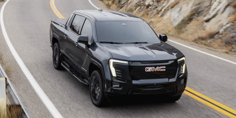 A dark gray GMC mid-sized pickup truck with glowing headlights speeding around a curve on an asphalt road.